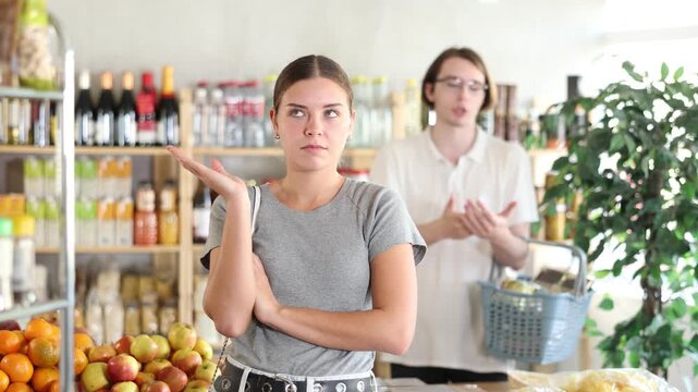 In shop guy stands behind wife, hurling abuse and indignation at her. In trading floor, sullen man stands with shop basket, swears with wife and listens to womans accusations. 