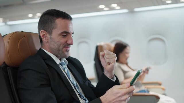 Businessman checks phone on plane. He reacts with excitement and satisfaction. Passenger sits calmly nearby. Ideal for travel, business
