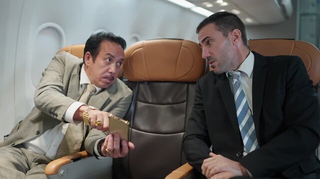 Two businessmen sit in airplane seats. One gestures with a phone while speaking. The other listens intently, leaning forward