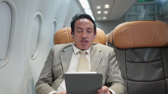 Businessman relaxes on airplane with tablet. He wears a suit and earpieces. He appears focused and comfortable. Ideal for travel or business themes