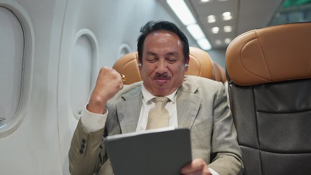 Man in suit sits airplane. He holds tablet, eyes closed, relaxed. He smiles, raises fist in joy. He celebrates success on flight. Ideal for business