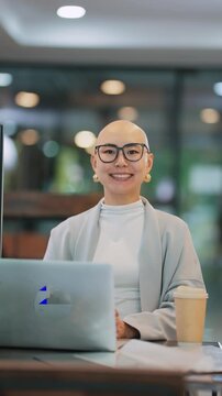 Professional woman with glasses smiles at camera. She sits at desk with laptop and coffee. Modern office background with soft lights