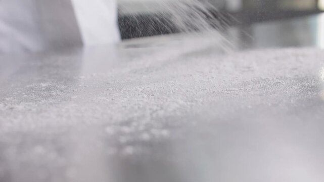 Flour is scattered across a clean metal kitchen work surface, creating a white dust cloud.