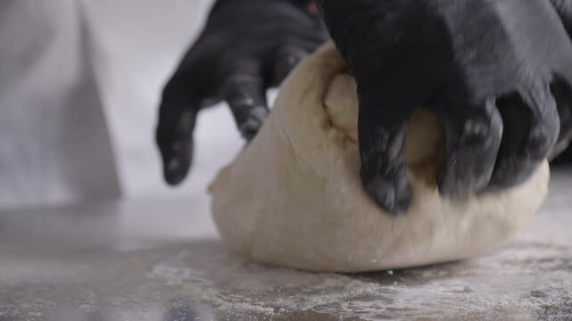 Black-gloved hands lift, fold and knead a large piece of dough on a floured metal counter.