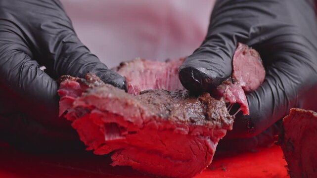 Black-gloved hands pull apart a piece of cooked meat, exposing its pink fibrous interior on a red cutting board.