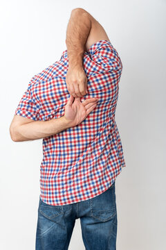 Person wearing a plaid shirt and jeans standing with both hands reaching behind the back, scratching upper back or shoulder blade area, indicating discomfort or an itch
