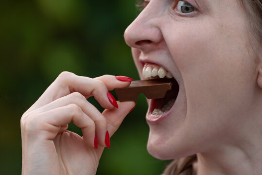 Young woman taking a bite of a delicious chocolate bar, her mouth wide open, craving the sweetness, and enjoying the indulgent moment with red fingernails
