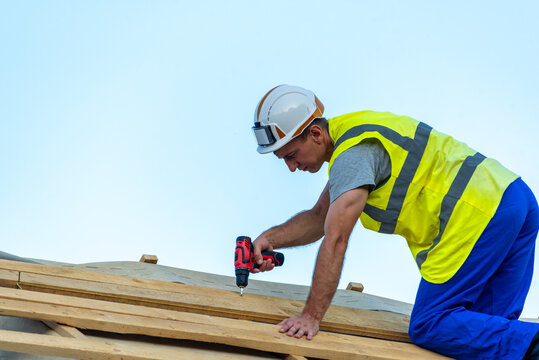 Construction worker wearing a hard hat and safety vest drilling wooden planks on a rooftop, building a new house under a clear blue sky, showing dedication and skilled labor