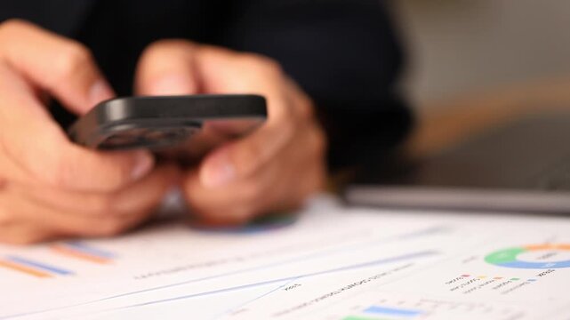 Close up of hands using a smartphone device reviewing financial data reports and business charts at an office desk, analyzing corporate growth and modern communication