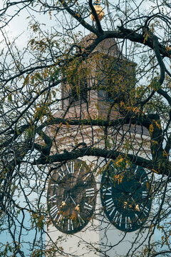 Clock tower of Petrovaradin Fortress in Novi Sad, Serbia, with two large black dials and Roman numerals seen through bare tree branches and autumn leaves against a pale blue sky.