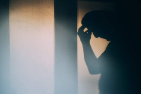 Blurred shadow of a worried figure touching their head in a dim room with soft window light, concept of depression, headache, loneliness and mental health