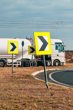 White tanker truck driving past yellow chevron road signs at a roundabout, with dry grass in the foreground, overhead power lines and a cloudy blue sky behind.