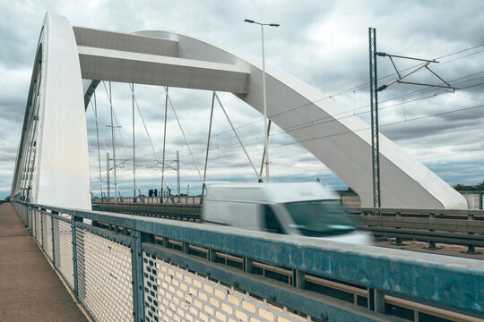 Motion blur of a delivery van crossing a city bridge with a large white concrete arch, suspension cables, lamp post and electric lines, seen from the pedestrian walkway.