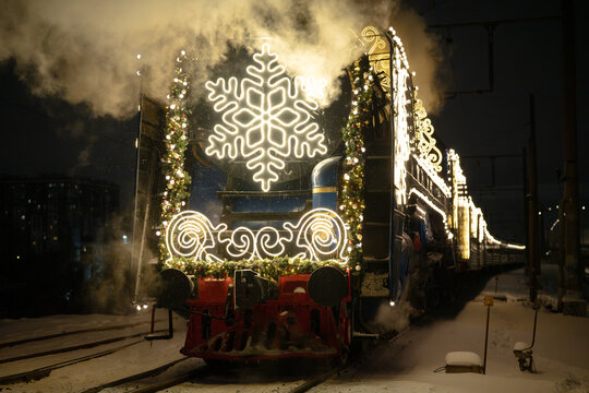 Decorated Christmas steam locomotive travels on snowy railway during frosty winter morning. Illuminated snowflake, garland and festive lights cover retro engine pulling holiday passenger carriage.