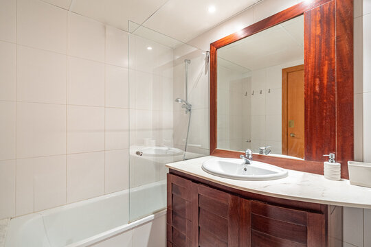 Modern bathroom interior with wooden vanity, large mirror, glass shower, white bathtub, chrome fixtures, bright recessed lighting, and minimalist contemporary design