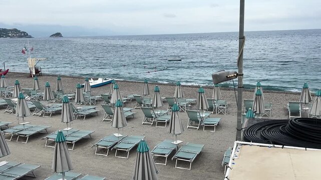 Peaceful pebble beach in Noli, Italy, featuring rows of turquoise sun loungers, closed umbrellas, calm Ligurian Sea waters, and a distant rocky island under cloudy skies.