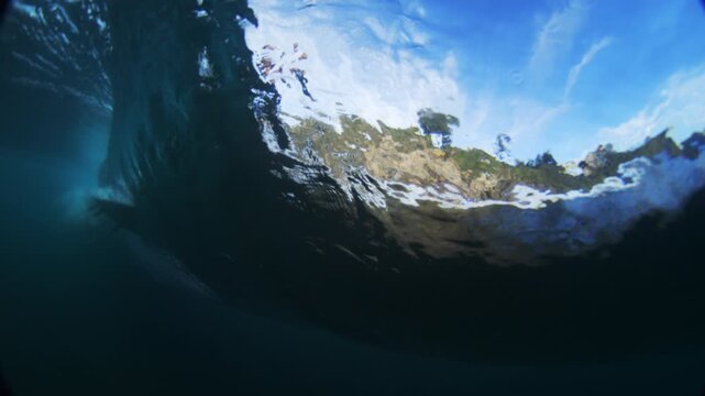 Blue ocean wave arches overhead, midday sky and rocky shore through the water surface, cliff edge and surfboard fin streaks