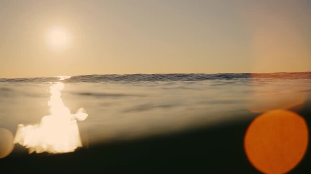 Warm sunset glows above ocean swells, golden reflection and large orange flare in slow motion, tranquil surface abstract