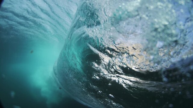 Sunlight shines through curling turquoise ocean water, white bubbles in close underwater view, backside rearview of barreling wave
