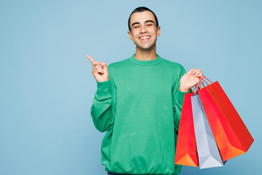 Young man wear green casual clothes hold paper package bags after shopping point thumb finger back aside on blank area isolated on plain pastel blue cyan background. Black Friday sale buy day concept.