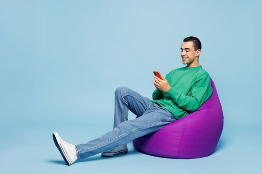 Full body young middle eastern man he wear green shirt casual clothes sit in bag chair hold in hand use mobile cell phone isolated on plain pastel light blue cyan background studio. Lifestyle concept.