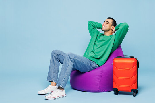 Traveler smiling happy man wear casual clothes sit in bag chair near suitcase bag isolated on plain blue background. Tourist travel abroad in free time rest getaway. Air flight trip journey concept