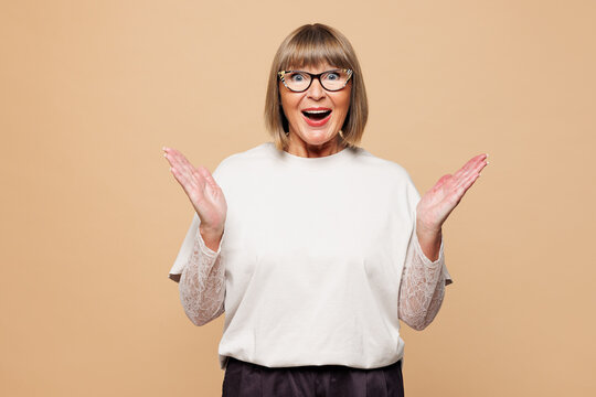 Elderly happy shocked surprised excited overjoyed blonde woman 50s years old she wear white shirt casual clothes spread hands look camera isolated on plain pastel beige background. Lifestyle concept.