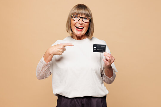Elderly blonde woman 50s years old wear white shirt casual clothes hold in hand point finger on mock up of credit bank card isolated on plain pastel light beige background studio. Lifestyle concept.