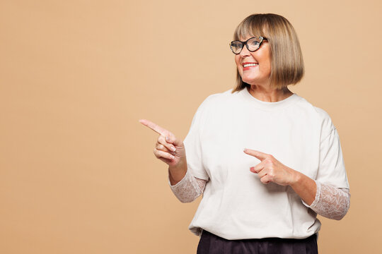 Elderly smiling happy blonde woman 50s years old she wear white shirt casual clothes point index finger aside area isolated on plain pastel light beige background studio portrait. Lifestyle concept