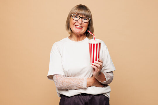 Elderly smiling happy blonde woman 50s years old she wear white shirt casual clothes hold in hand cup of soda pop cola fizzy water isolated on plain pastel light beige background. Lifestyle concept.