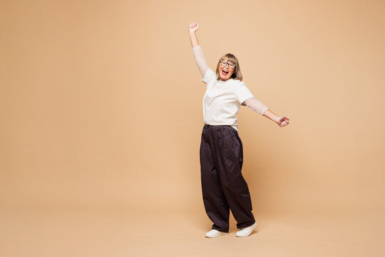 Full body smiling happy elderly blonde woman 50s years old she wear white shirt casual clothes do winner gesture clench fist say yes isolated on plain pastel light beige background. Lifestyle concept.