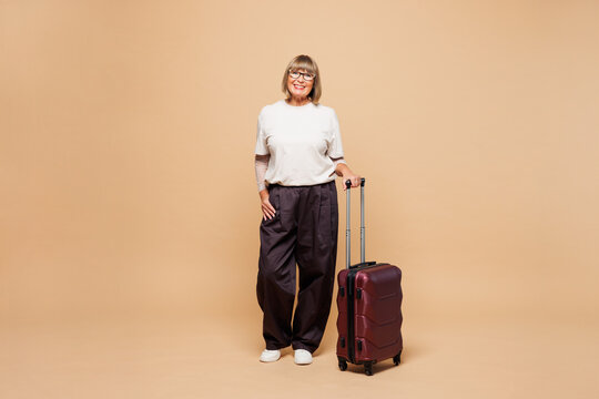 Traveler happy elderly woman she wear white casual clothes hold bag suitcase isolated on plain beige background. Tourist travel abroad in free spare time rest getaway. Air flight trip journey concept