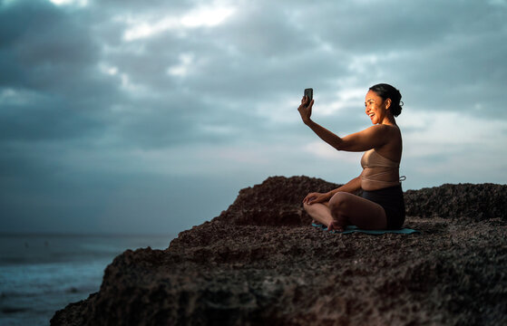 Asian woman taking a smartphone selfie while sitting in yoga pose on an ocean cliff