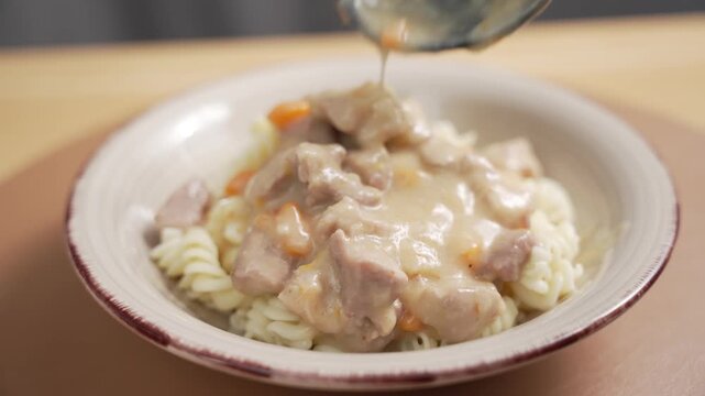 A ladle places creamy meat stew with carrot pieces on top of fusilli pasta in a ceramic bowl on a table.