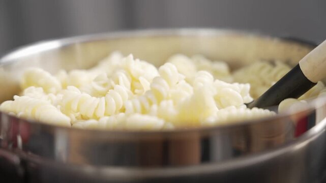 Cooked fusilli pasta falls into a stainless steel pot, then a black pasta server with a wooden handle stirs and lifts it.