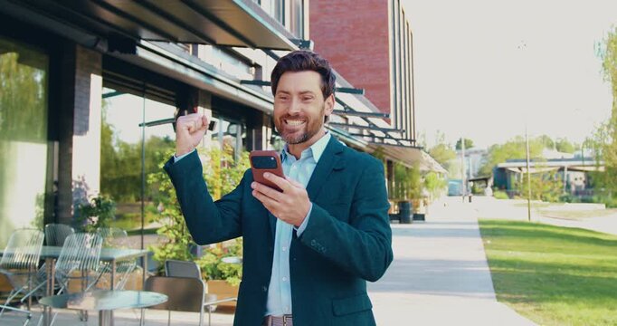 Businessman standing on a city street, looking at his smartphone and reacting with excitement to good news on the screen. He smiles, celebrates and gestures with his hands while reading the message.