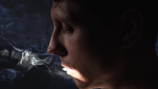 Extreme close-up of a man finishing a cigarette with lowered eyes, conveying nicotine dependence and loss of control, dark cinematic low-key lighting and visible smoke create an intimate mood