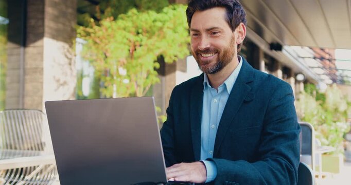 Professional businessman in business attire sitting at an outdoor table, working on a laptop and smiling.