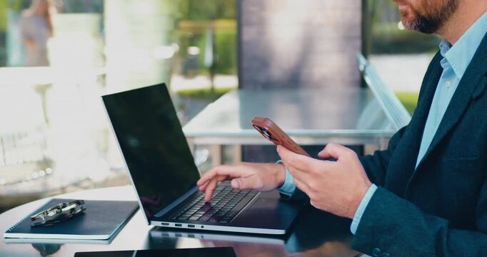 Businessman in a formal suit browsing work-related content on a smartphone and typing on a laptop at an outdoor table.