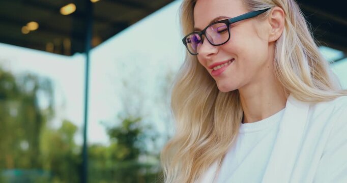 Close-up of smiling blonde woman wearing glasses looking at laptop screen outdoors