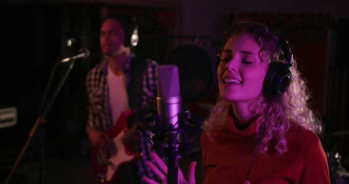 Blonde female singer records a song with a guitarist in a studio.