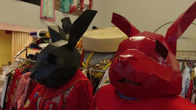Two people in sculptural rabbit masks browse thrift shop clothing rack, dressed in vivid red and patterned garments. Unusual costumes give this playful retail moment artistic edge. Halloween