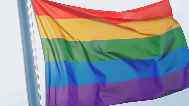 Rainbow pride flag waving from a metal flagpole beneath a bright open sky. Lgbtq awareness campaigns, diversity and inclusion advertising, and human rights communications.