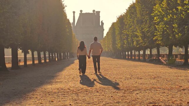A couple strolls along a tree-lined garden path in Paris France, their long shadows stretching across the ground in warm evening light.