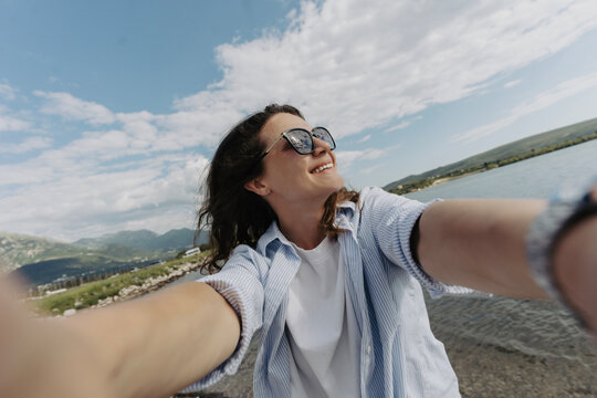 Smiling young woman in sunglasses taking selfie on sea shore in windy weather