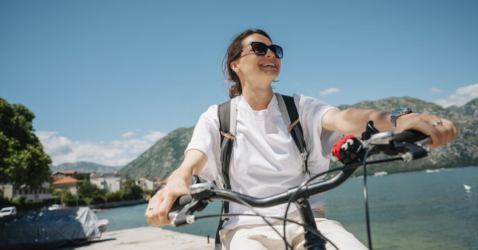 Smiling young Caucasian woman riding city cruiser bicycle along scenic Kotor Bay in Montenegro