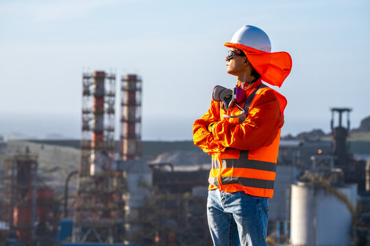 Industrial worker wearing high-visibility protective gear and respirator, surveying a refinery