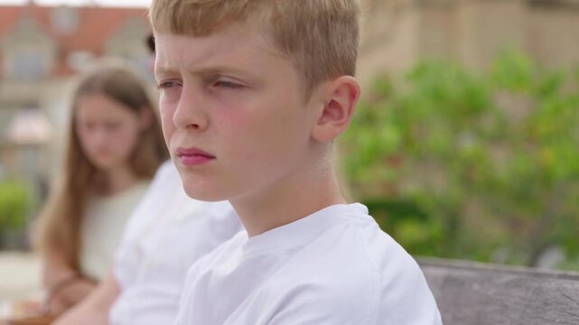 A sad boy looks at the camera slow motion in the background his family mother and sister are sitting on a bench