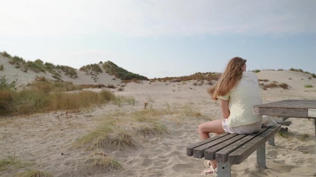 Girl sitting on a wooden bench among coastal sand dunes adjusting shoes gets up and walks away rear view