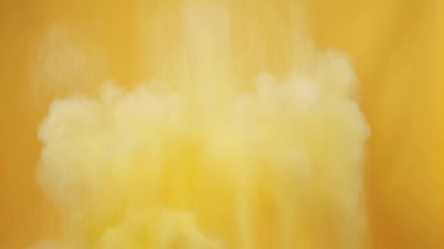 Pale yellow plumes billow and spread across golden background, forming soft cloudlike shapes among fine bright particles in luminous abstract composition with warm amber tones